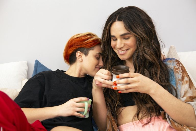 A transmasculine gender-nonconforming person and transfeminine nonbinary person drinking coffee in bed