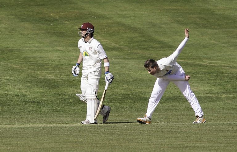 A cricket match action shot showing a bowler mid-delivery on a grass field while a batsman stands nearby