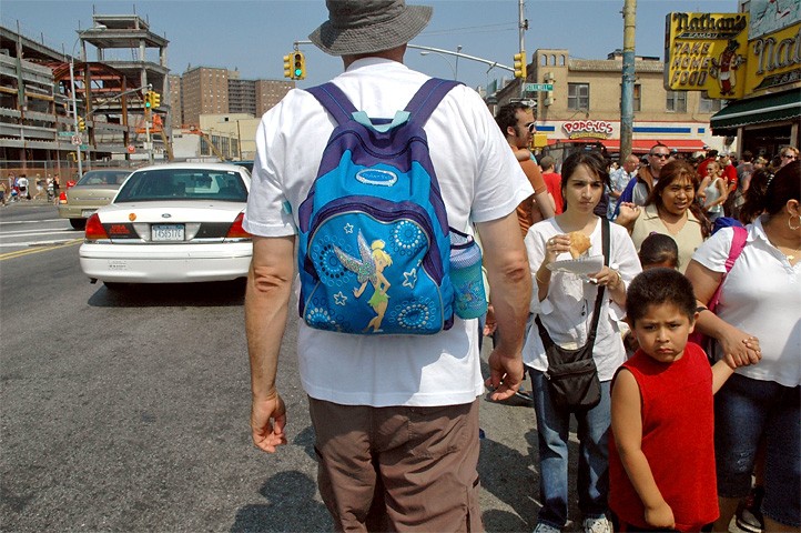 A man from behind wearing a small, bright blue Tinker Bell kids' backpack while walking on a crowded city street.