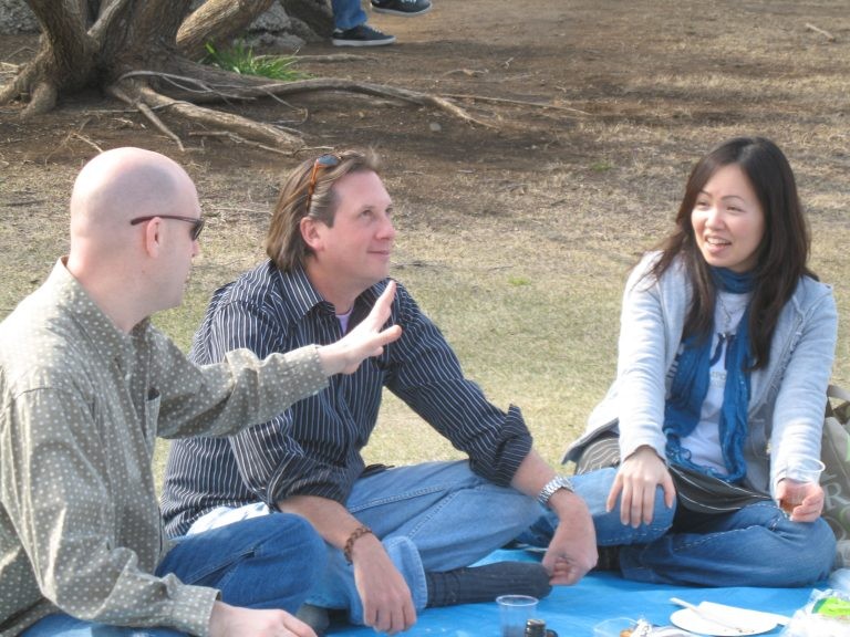 Three people sitting cross-legged on a blue picnic tarp outdoors, with one man gesturing with an open hand while speaking.
