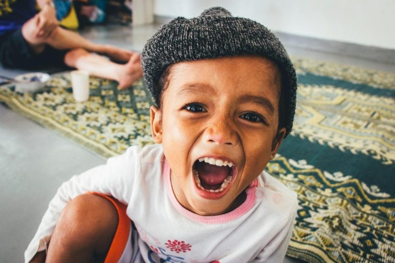 A close-up shot of a young child wearing a gray knit beanie cap, looking at the camera with a wide, open-mouthed smile.