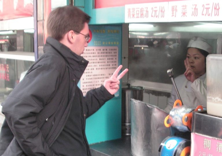 A man ordering food at an outdoor stall, making a "two" hand gesture to the worker behind the counter.