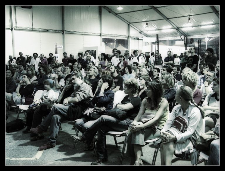 A large audience sitting in rows of chairs inside a tented event space, focused intently on a presentation.