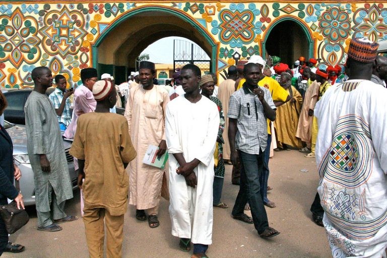 A crowd gathered outside a palace building adorned with vibrant, colorful traditional Hausa geometric architectural patterns.
