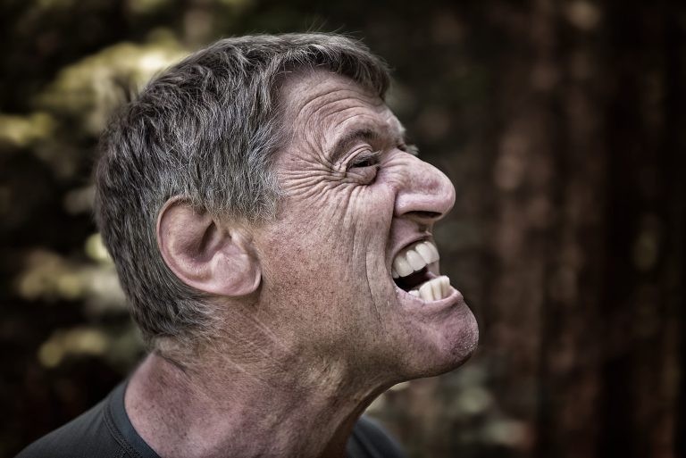 A close-up side profile portrait of a man grimacing intensely with a wide, bared-teeth facial expression.