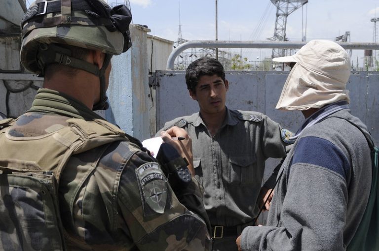 An ISAF soldier with a NATO patch takes notes while speaking with a uniformed man and a civilian outdoors.