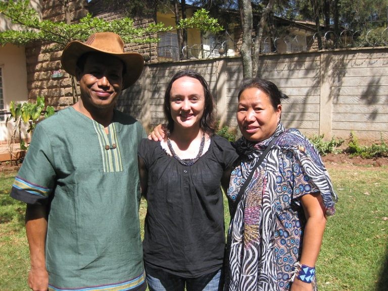 Three smiling people stand together on a lawn, including a man in a brown cowboy hat and green tunic.