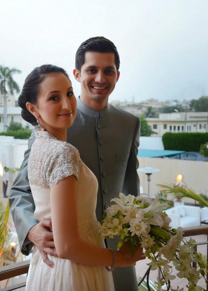 Smiling bride in an elegant white dress holding a lily bouquet while her groom in a grey tunic embraces her.
