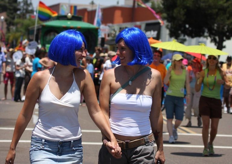 Two smiling women wearing bright blue wigs hold hands as they walk outdoors in a crowded, festive parade.