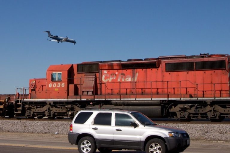 An SUV drives on a road in the foreground with a large red CP Rail train engine behind it and an airplane flying above.