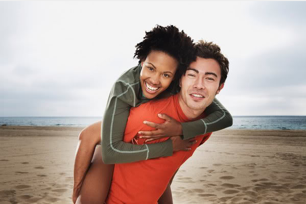 A smiling man gives a woman a piggyback ride on a sandy beach with the ocean visible in the background.