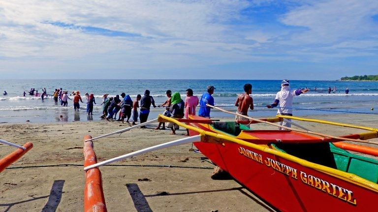 A group of people pulls a fishing net from the sea onto a sandy beach next to a red outrigger boat.