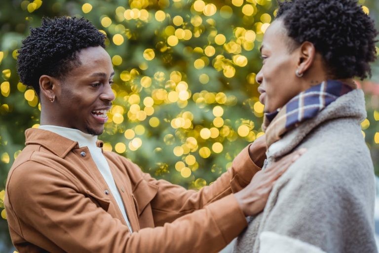 A smiling man in a brown jacket looks affectionately at his partner in front of blurred yellow holiday lights.