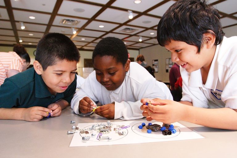 Three boys are smiling as they sort small metal screws and blue plastic pieces on a table during a classroom activity.