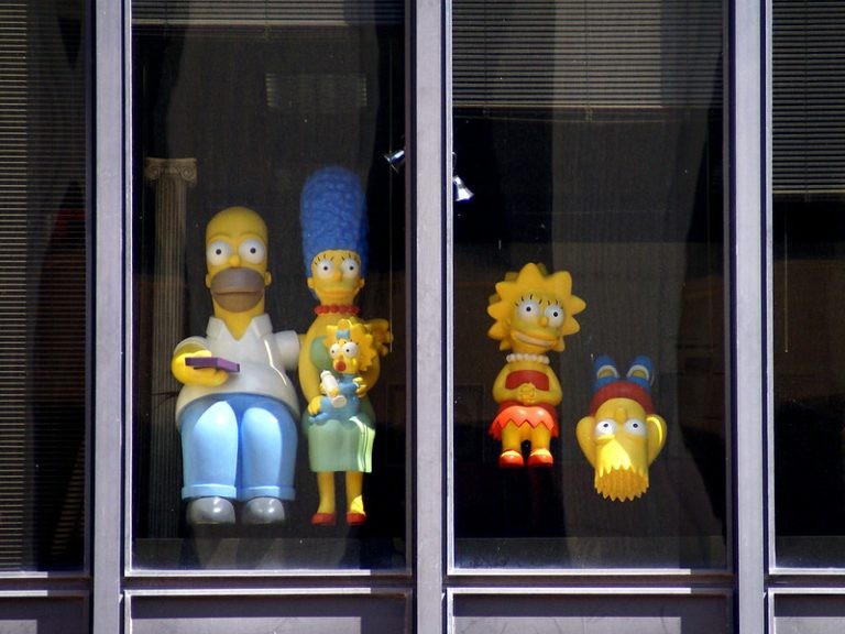Figures of the Simpsons family displayed in a window, featuring Homer, Marge, Maggie, Lisa, and Bart upside down.