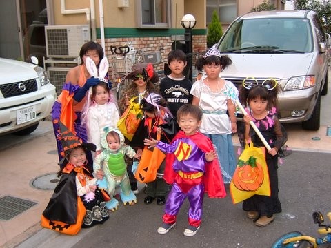 A group of young children posing outside in various Halloween costumes, including a witch and a superhero.