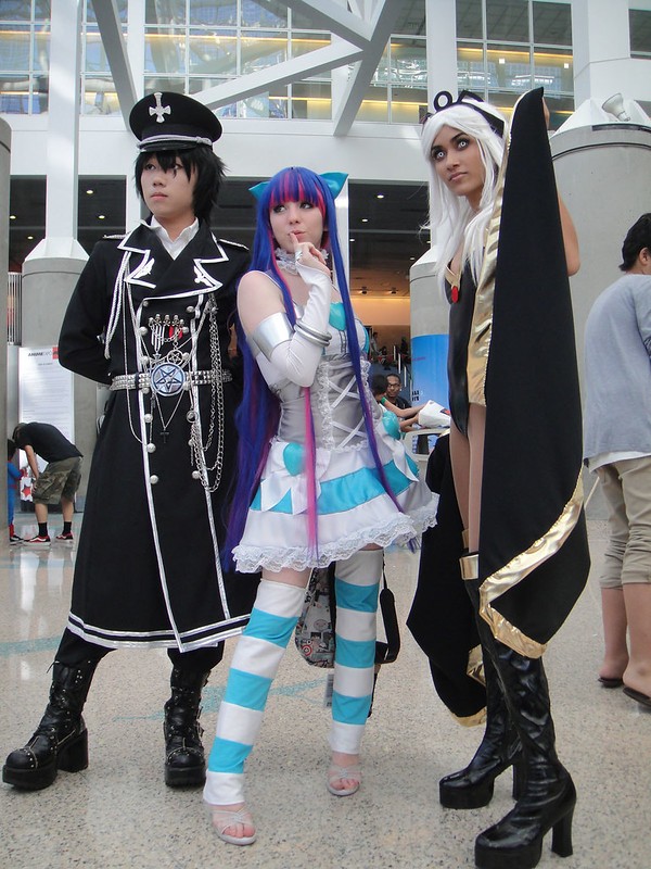 Three cosplayers posing in detailed anime and comic book character costumes at an indoor convention venue.