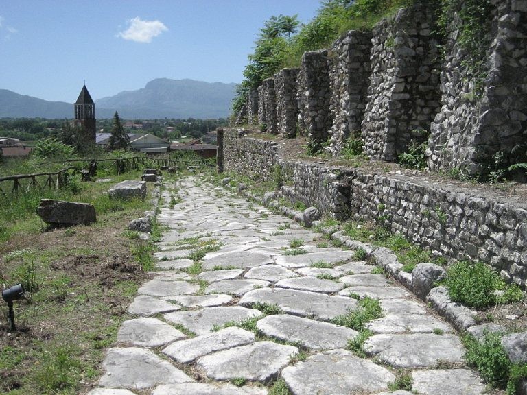An ancient, stone-paved Roman road runs alongside stone ruins on a grassy hillside overlooking a distant town and mountains.