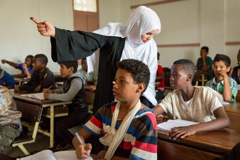 A teacher in a hijab gestures to guide a lesson for elementary school students writing at wooden desks in class