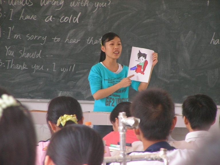 A student teacher in China holds a flashcard of a sick boy with conversation prompts written on a chalkboard