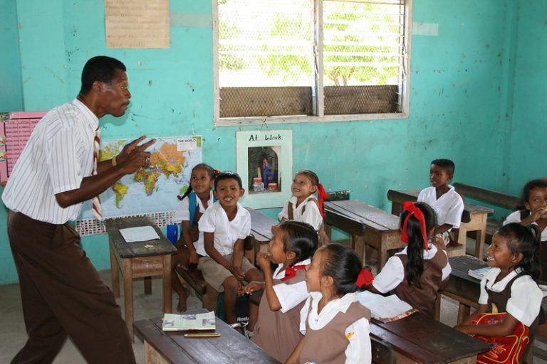 A teacher is gesturing at the chalkboard during a lesson for elementary students sitting at wooden desks in a classroom.