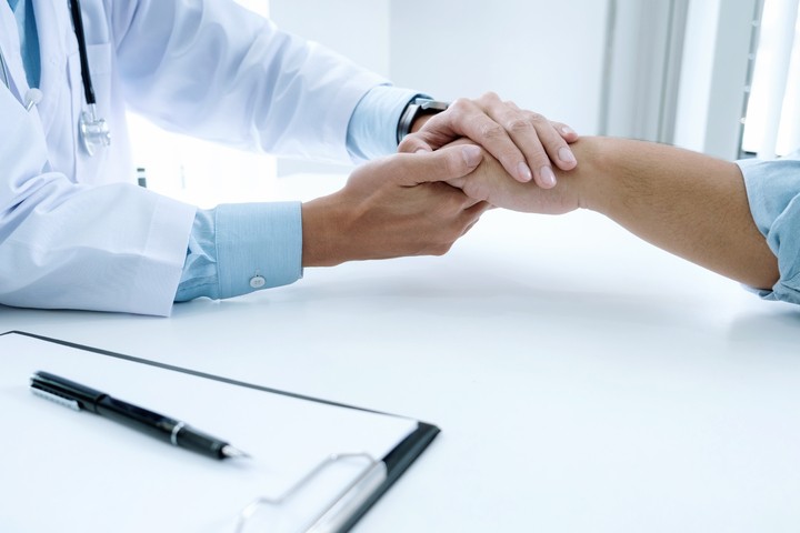 A doctor wearing a white lab coat comforts a patient by holding their hand over a desk with a clipboard