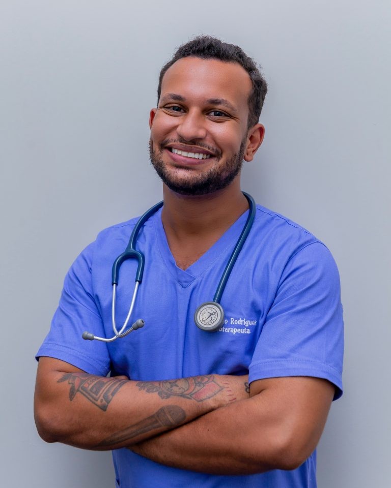 Smiling male healthcare professional wearing blue scrubs and a stethoscope around his neck with arms crossed.
