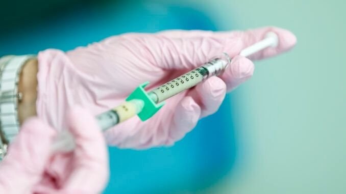 Gloved hands connecting two syringes with a green adapter to transfer or mix a liquid medication in a clinic.