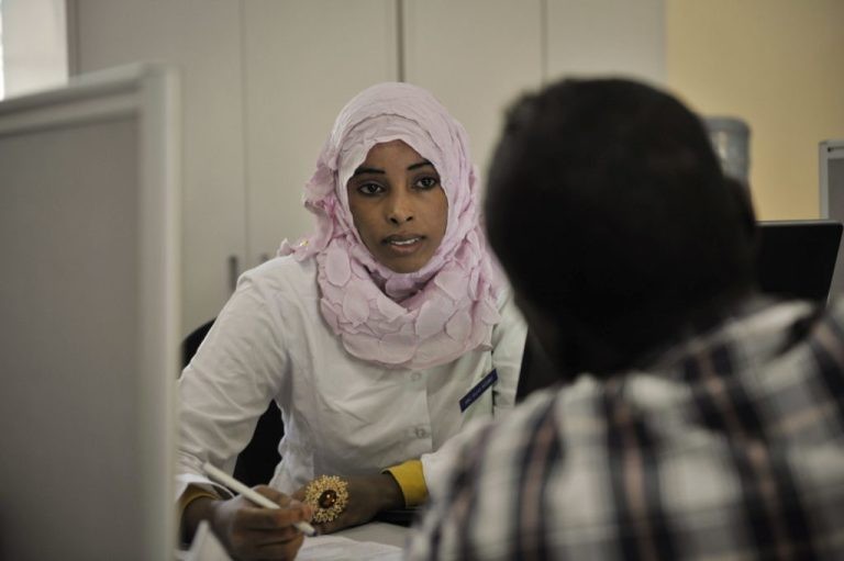 A professional woman in a hijab and lab coat speaking with a person at a desk in a clinical setting.