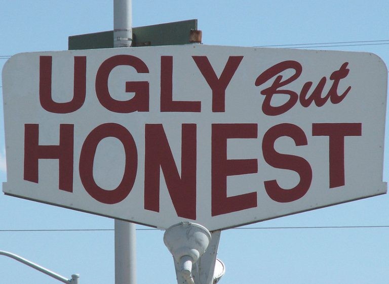 A white roadside sign featuring the words "UGLY But HONEST" painted in large, bold red letters against a clear blue sky.
