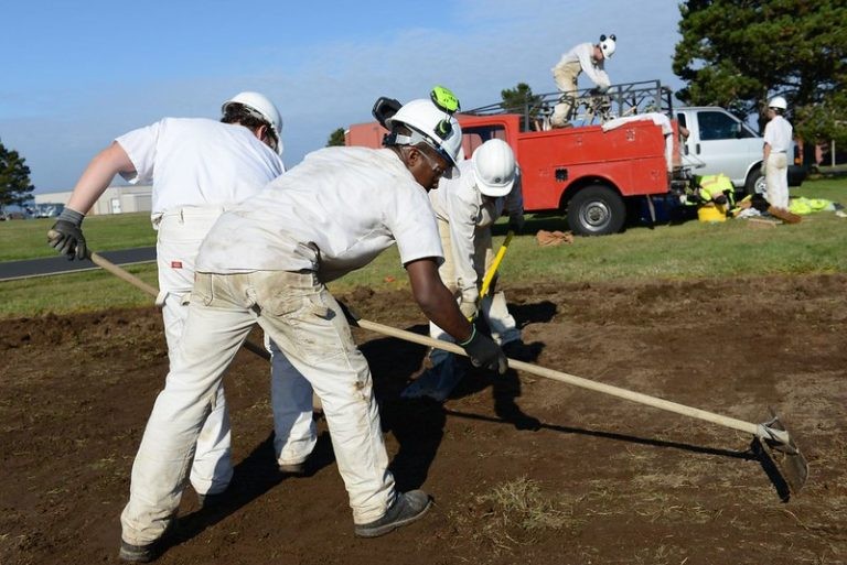A group of workers in white uniforms and hard hats clear a dirt field with hand tools near a red truck.