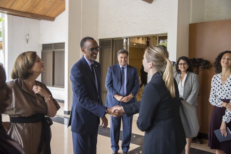 A tall man in a dark suit shakes hands with a blonde woman in a black blazer as several onlookers smile in an indoor lobby.