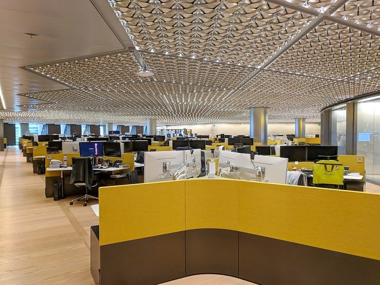 Spacious, open-plan office with rows of desks, yellow dividers, computer monitors, and a unique textured metallic ceiling.