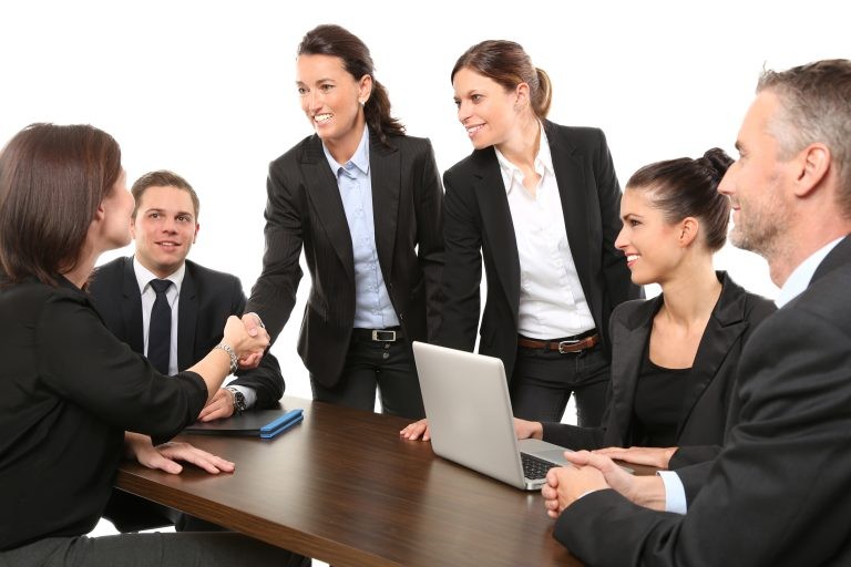 A group of business professionals in suits smiles during a meeting around a conference table while two women shake hands.