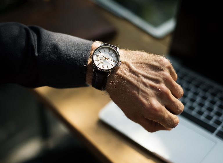 Close-up view of a man's arm and wrist shows a classic round watch with a leather strap as he checks the time over a laptop