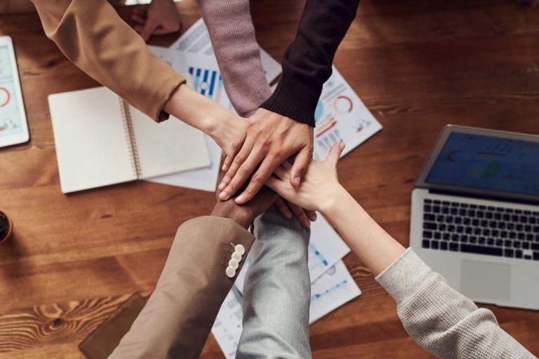 An overhead view of a diverse group of business colleagues stacking their hands together over a wooden table with laptops.