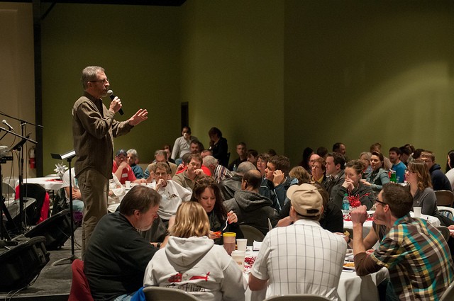A speaker stands with a microphone, addressing a large group of people seated around tables at an event.