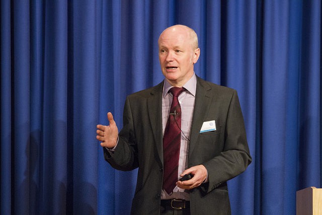 A man in a suit is gesturing with his hand while speaking in front of a blue stage curtain
