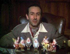 Walk Disney sitting in an office chair. On the desk in front of him are the 7 dwarves as dolls