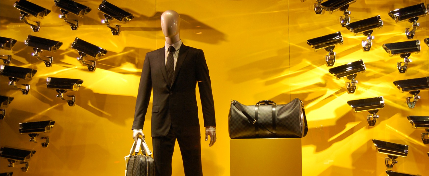 A suited mannequin surrounded by many security cameras, on a bright yellow background with two designer bags in view.
