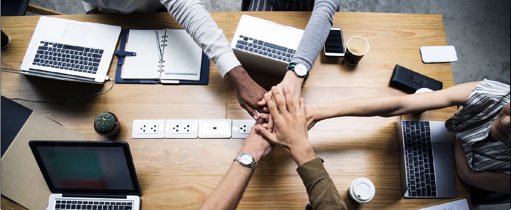 Overhead shot of diverse hands in a team huddle; laptops, a cactus, and other office objects surround it.