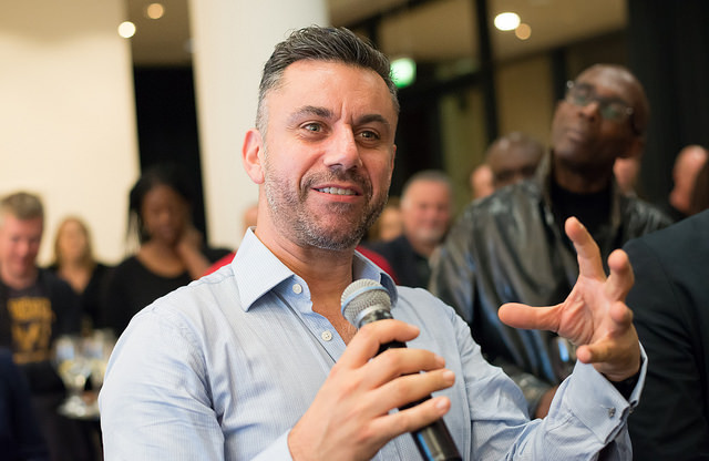 A man holding a microphone and gesturing with his hand while speaking to an audience at an indoor event.