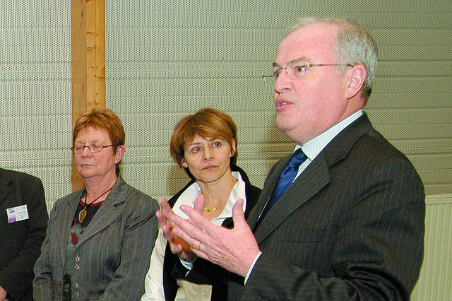 Christian Pierret, in a suit and glasses, gestures while speaking, with two women listening in the background.
