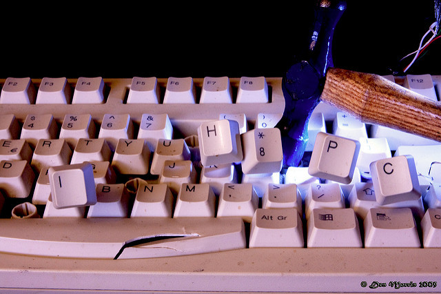 A computer keyboard is being smashed with a hammer, causing several keycaps to pop off.