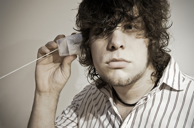 A young man with dark hair holds a white plastic cup attached to a string to his ear, listening intently.