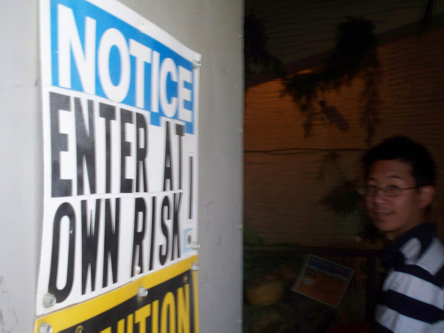 Close-up of a sign reading Notice: Enter at Your Own Risk with a man wearing glasses standing in the background.