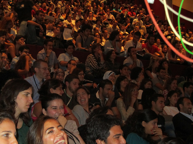 A large, crowded indoor auditorium filled with people sitting in rows of tiered seating and smiling.