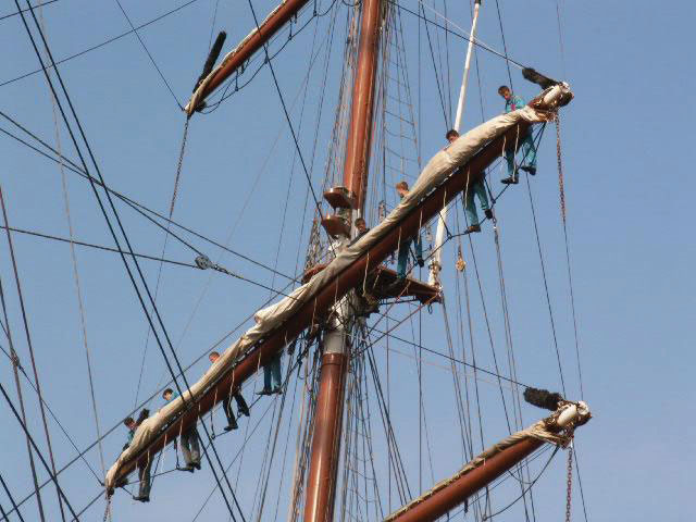 Several sailors are climbing up and balancing along the wooden yardarms of a tall sailing ship under a clear blue sky.