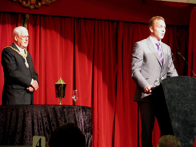 A man in a suit speaks from a podium on a stage with red curtains, while another man stands nearby, listening.