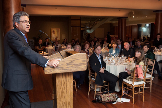 A man in a dark suit speaks at a wooden podium to a seated audience at a formal indoor event.